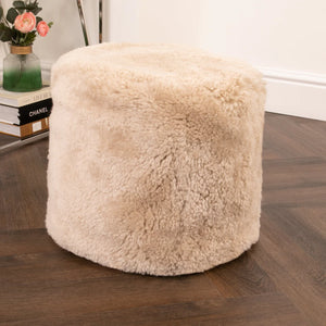 Beige fluffy ottoman on a wooden floor with books and a vase in the background.