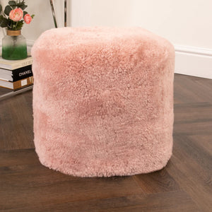 Pink fluffy ottoman on a wooden floor with books and a vase in the background.