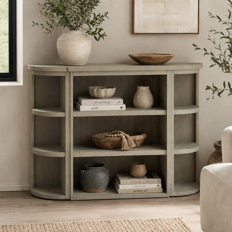 A curved wooden console shelf unit in a weathered grey finish, styled with books, ceramic vases, a bowl, and a potted plant in a bright living room setting.