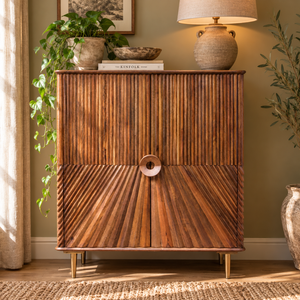 A mid-century modern wooden sideboard cabinet with vertically and diagonally slatted doors, gold legs, and a circular wooden handle, styled in a warm, sunlit room with plants and a lamp.