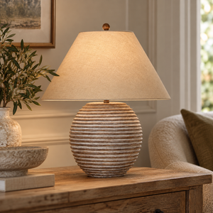 Saturn Table Lamp With Linen Shade lit on a wooden side table beside an armchair and olive branches