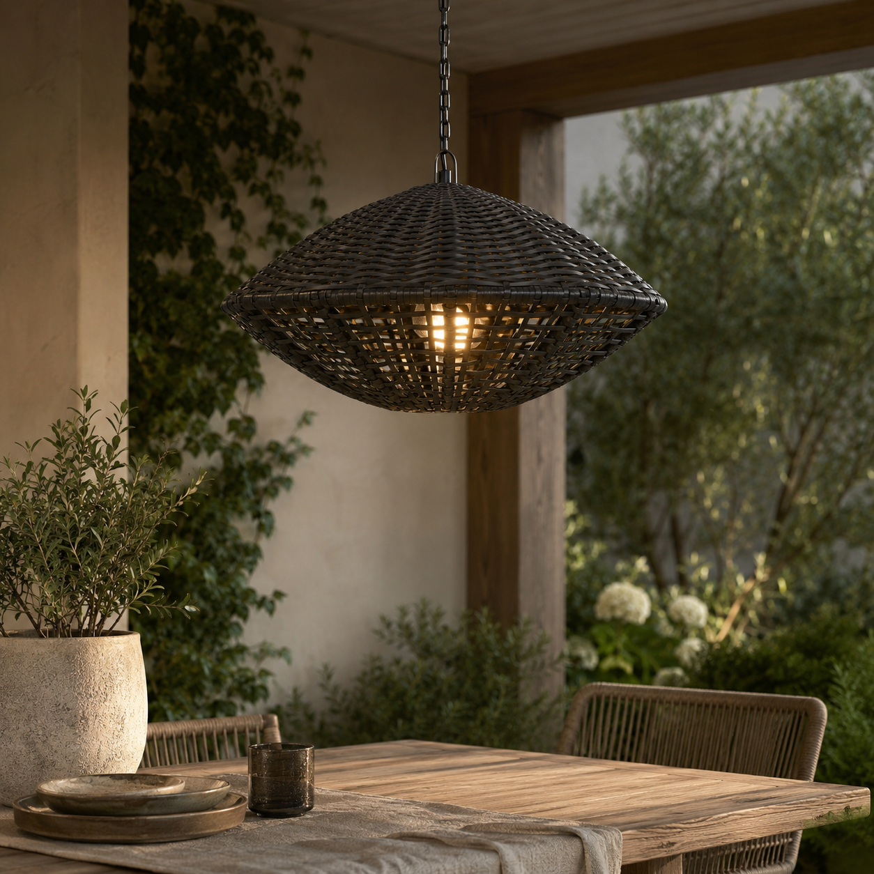 A dark woven pendant light hangs over a wooden dining table on a covered patio with plants and outdoor greenery in the background.