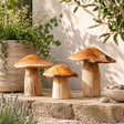 Three rustic wooden mushroom garden statues in different sizes, displayed on stone steps in a garden setting with potted plants and a textured wall.