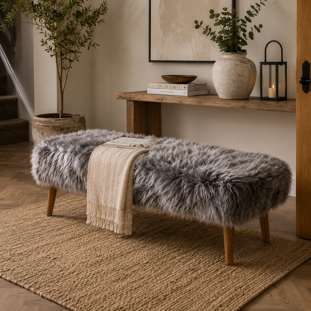 A grey shearling sheepskin bench with wooden legs sits on a woven jute rug in a styled entryway. A cream throw blanket is draped over the bench, and a console table with books, a ceramic vase, and a lantern is visible in the background.