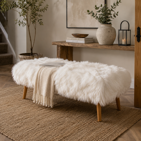 A white shearling sheepskin bench with light wood legs in a styled living room setting, featuring a cream throw blanket draped over the front, a woven rug, a wooden console table, and potted plants.