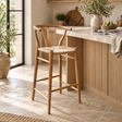 A light natural wood bar stool with a woven rope seat sits beside a stone-topped kitchen island in a bright, sunlit kitchen.
