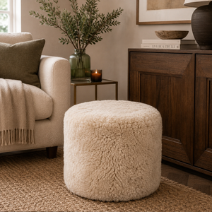 A round, fluffy cream-colored pouffe ottoman in a styled living room setting next to a beige armchair and a dark wood cabinet.