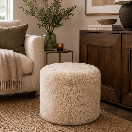 A round, fluffy cream-colored pouffe ottoman in a styled living room setting next to a beige armchair and a dark wood cabinet.