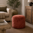 A round, textured terracotta pouffe ottoman in a cosy, rustic living room setting next to a beige armchair and wooden furniture.
