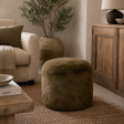 An olive green shaggy sheepskin cylindrical pouf ottoman in a cosy, modern living room setting next to a cream armchair and a wooden cabinet.