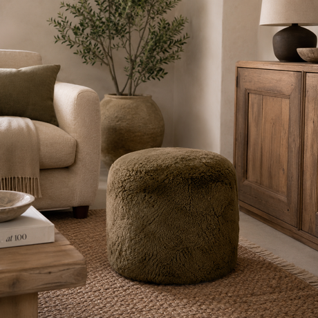 An olive green shaggy sheepskin cylindrical pouf ottoman in a cosy, modern living room setting next to a cream armchair and a wooden cabinet.