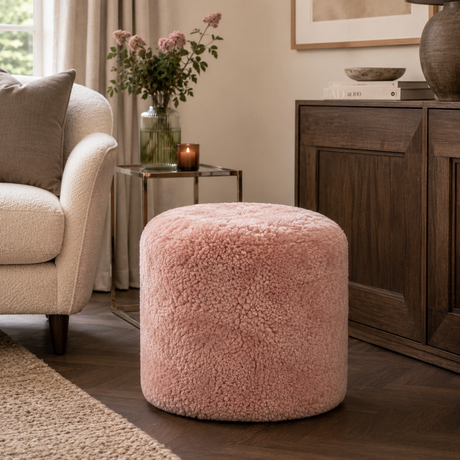 A round, plush pouffe ottoman in a dusty pink shorn sheepskin texture, styled in a cosy living room next to a cream armchair and a dark wood sideboard.