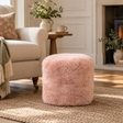 Blush pink sheepskin pouffe in a cosy neutral living room, styled beside a cream armchair on a natural woven rug.