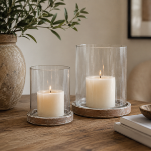 Two clear glass cylinder candle holders with lit white pillar candles, displayed on round stone-like coasters on a wooden table in a warm, styled interior setting.