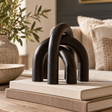 Silva Black Wood Arch Sculpture Ornament on a stack of linen-covered books on a wooden table, with a cream sofa and olive branches behind