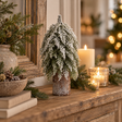 Small snowy fir tree on a tall bark-covered log on a mantel with lit candles and pine greenery