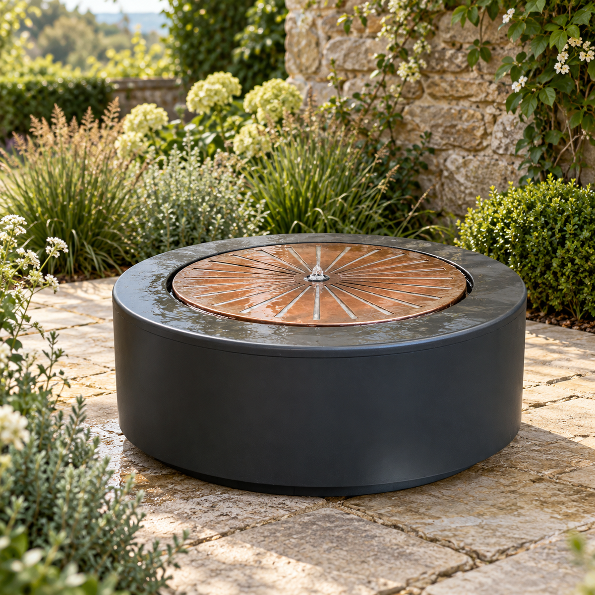 Solis Large Water Feature in Charcoal & Aged Copper on a sunlit stone terrace, surrounded by hydrangeas, grasses and a stone wall