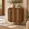 A mid-century modern wooden sideboard cabinet with two doors featuring chevron wood inlay patterns, standing in a warm, sunlit room with stone tile flooring and decorative plants on top.