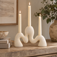 Soren Cream Organic Candle Holder with three lit cream pillar candles in its sculptural wavy, speckled form, set on a rustic wooden table in a warm, styled interior.