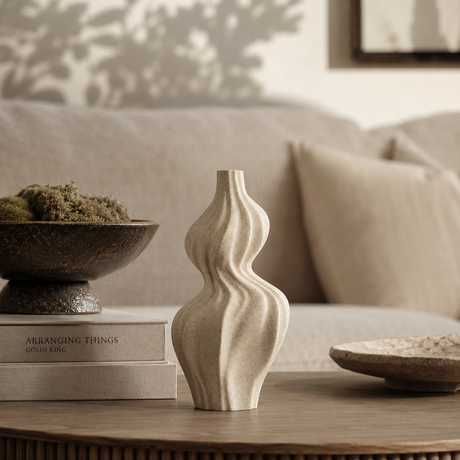 A beige ceramic vase with a sculptural, wavy ribbed design sits on a wooden table in a cozy, modern living room setting next to a stack of books and a dark bowl.