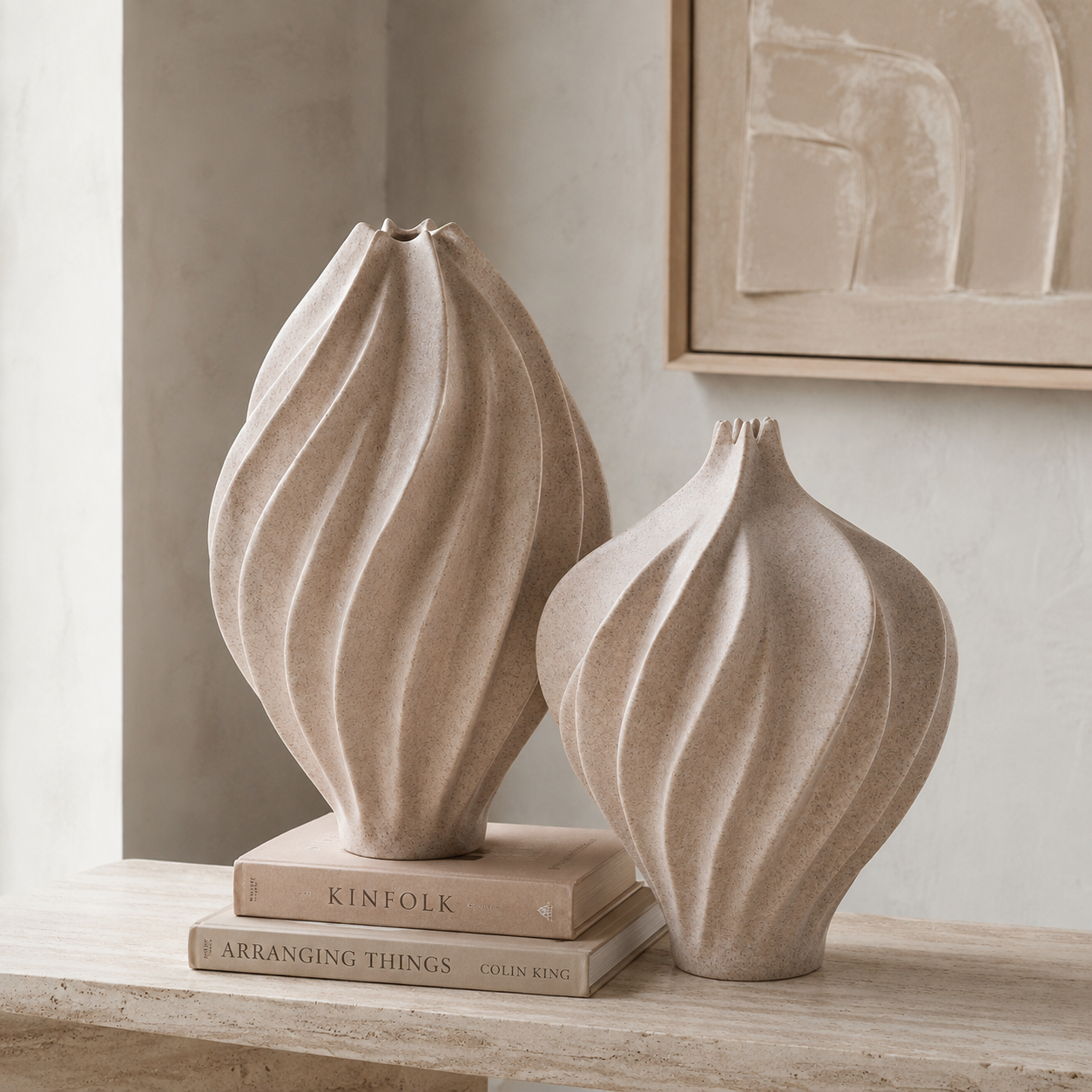 Two ribbed ceramic vases in a warm beige tone, one standing on stacked books and the other beside it, displayed in a softly lit interior setting.