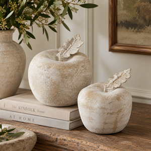 Two Stone Apples in different sizes on a wooden sideboard, the larger resting on a stack of books, beside a stoneware vase of olive branches