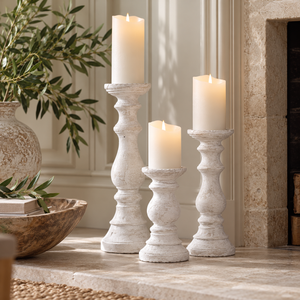 Three Stone Candle Holders in different heights with cream pillar candles, grouped on a stone hearth beside a fireplace and a vase of olive branches