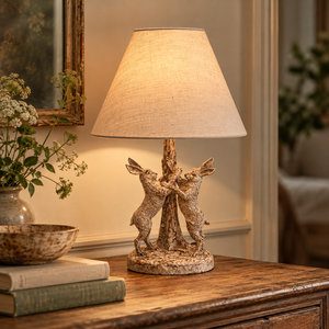 Stonewashed Marching Hares Lamp With Linen Shade lit on a wooden chest beside wildflowers and books