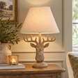 Stonewashed Stag Head Table Lamp With Linen Shade lit on a wooden side table beside a candle and greenery