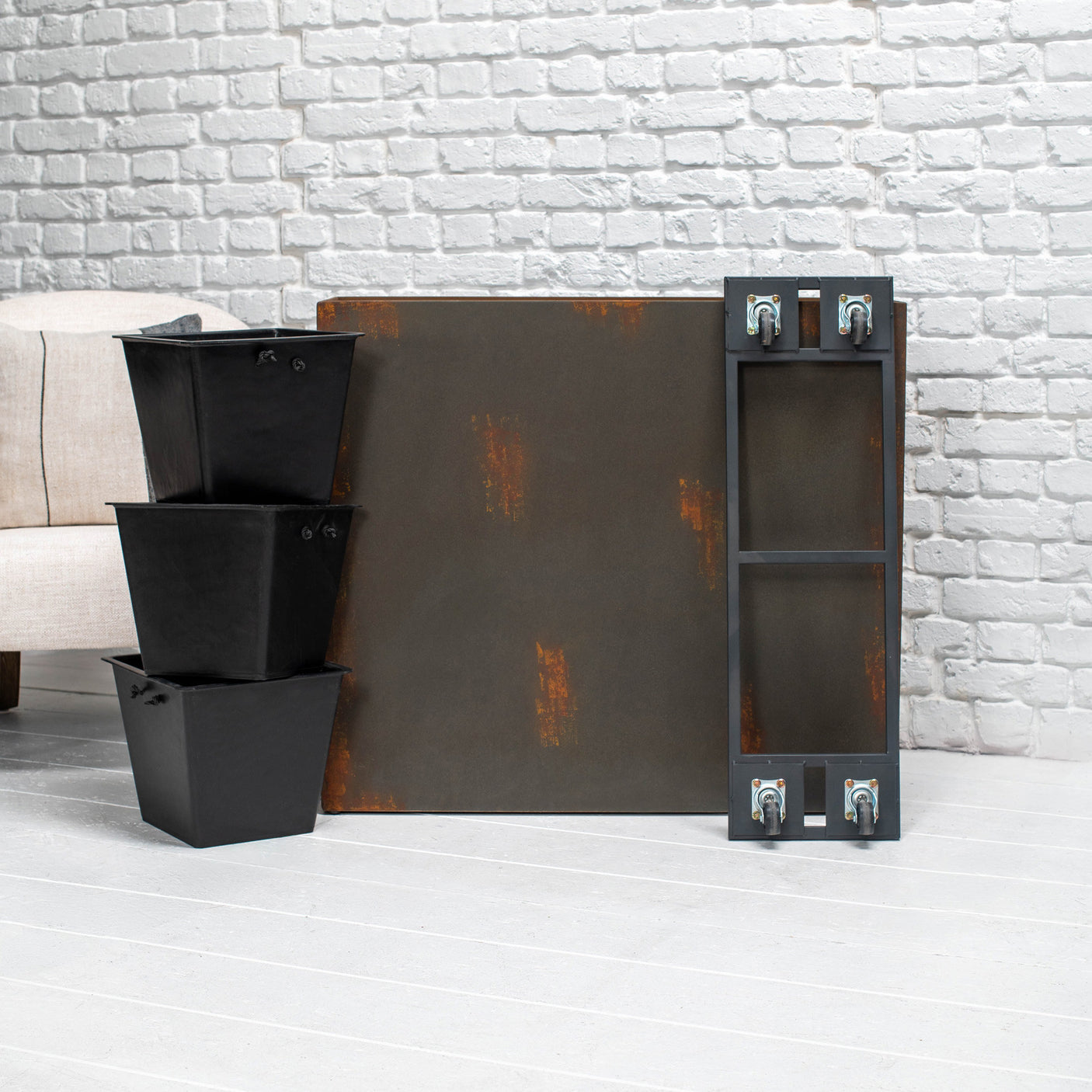 A rustic industrial-style rectangular metal planter with distressed rust patches and a tall narrow side frame with wheels, shown next to three stacked black storage bins against a white brick wall.