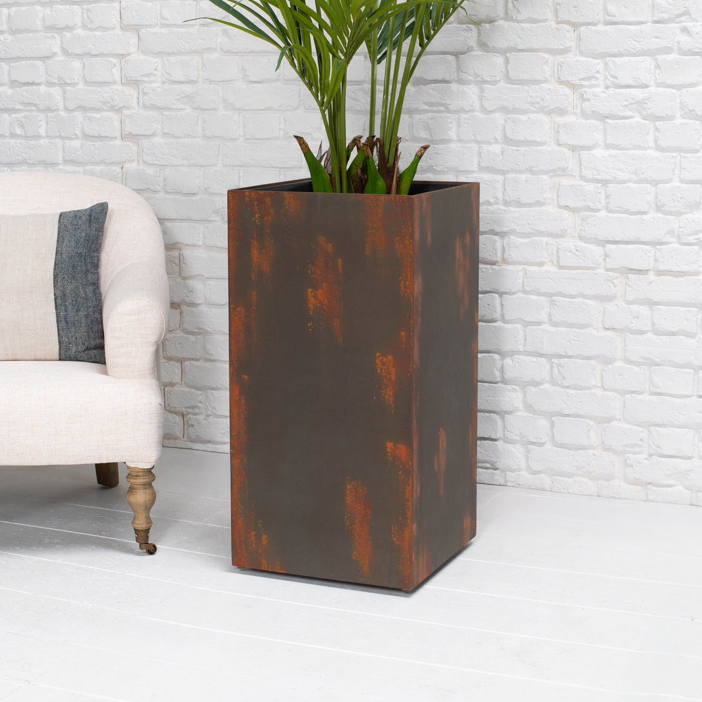 A tall rectangular metal planter with a distressed rust-brown finish, holding a green palm plant, set against a white brick wall and white floor, next to a light-colored armchair.