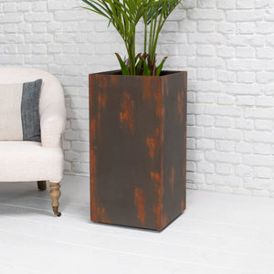 A tall rectangular metal planter with a distressed rust-brown finish, holding a green palm plant, set against a white brick wall and white floor, next to a light-colored armchair.