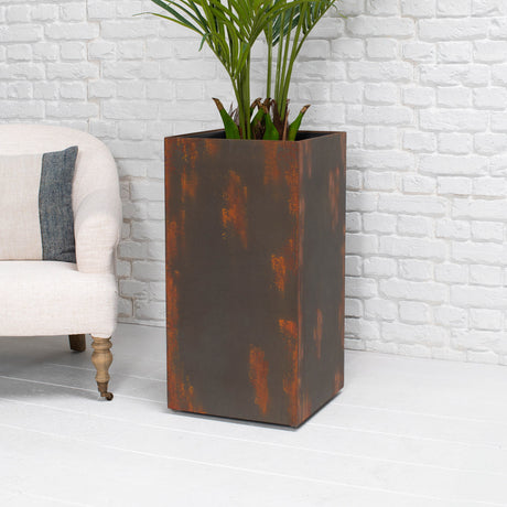 A tall rectangular metal planter with a distressed rust-brown finish, holding a green palm plant, set against a white brick wall and white floor, next to a light-colored armchair.