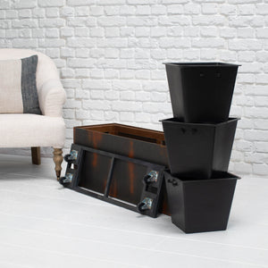 A black metal storage planter box with wheels, shown alongside three nested black square planters on a white brick floor next to a light-colored armchair.