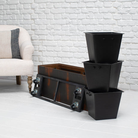 A black metal storage planter box with wheels, shown alongside three nested black square planters on a white brick floor next to a light-colored armchair.
