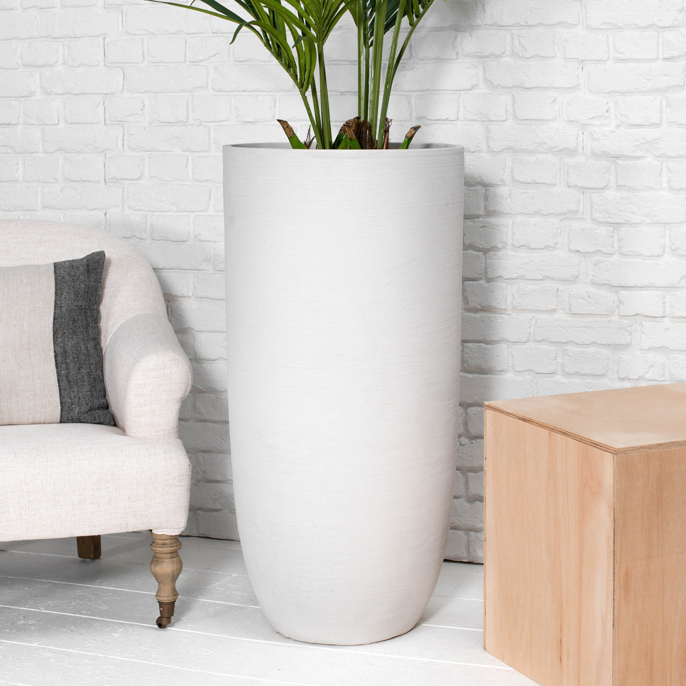 A tall, cylindrical matte white planter with a green plant, set against a white brick wall in a styled room with a beige armchair and a light wood side table.