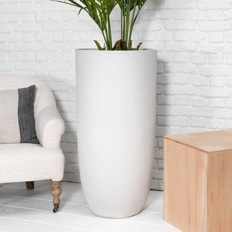 A tall, cylindrical matte white planter with a green plant, set against a white brick wall in a styled room with a beige armchair and a light wood side table.