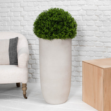 A tall cylindrical matte beige planter holding a dense, spherical green topiary tree, styled in a living room setting with a white brick wall, a light-colored armchair, and a wooden side table.