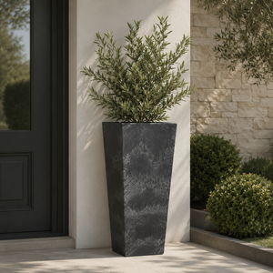 A tall black artstone planter in an outdoor setting against a cream wall with plants and a black front door