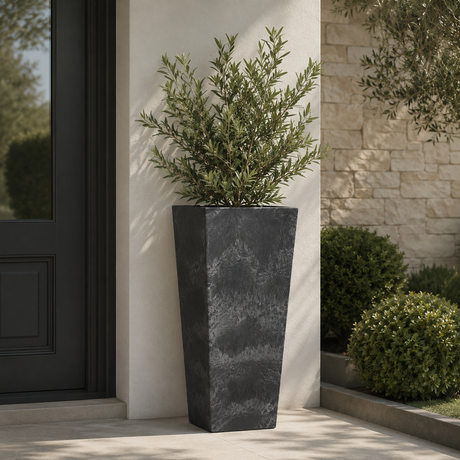 A tall black artstone planter in an outdoor setting against a cream wall with plants and a black front door