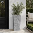 A tall grey outdoor planter against a white wall in an outdoor setting beside a black front door 