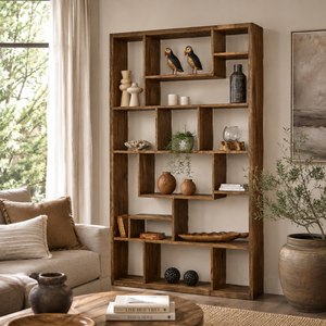 A tall, multi-compartment wooden bookcase with a rustic medium-brown finish, styled in a cozy living room setting next to a beige sofa and potted plants.