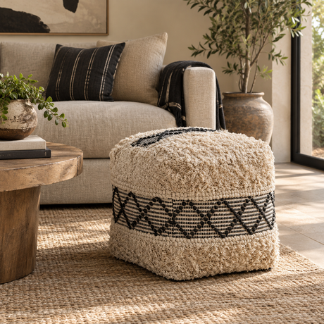 A square shaggy pouffe in cream with a black geometric pattern band, placed in a styled living room next to a beige sofa and a wooden coffee table.