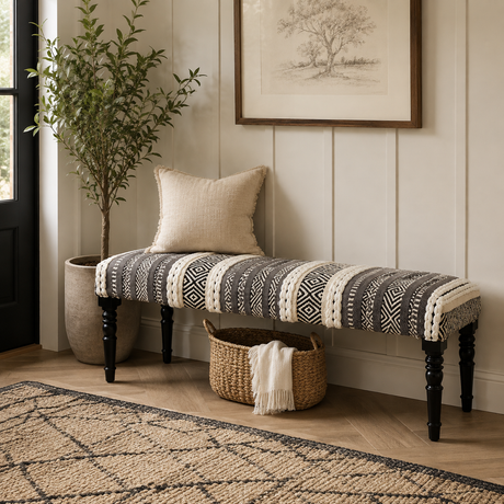 A long upholstered bench with a black-and-white geometric patterned seat cushion and dark turned legs, styled in a home entryway with a beige pillow, woven basket, plant, framed wall art, and wood flooring.