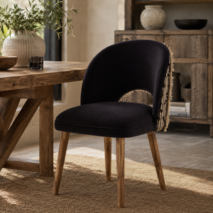 A black velvet dining chair with light brown wooden legs and a gold beaded fringe detail on the backrest, positioned next to a rustic wooden dining table in a warm, modern dining room setting.