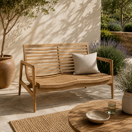 A two-seater wooden outdoor lounge bench with a slatted back and seat, styled on a patio with a neutral throw pillow, potted plants, and a stone wall background.