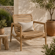 Teak outdoor lounge chair with a slatted back and seat, styled with a neutral cushion on a sunlit patio surrounded by plants and terracotta pots.