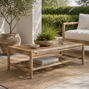 Natural wood outdoor coffee table with a slatted top and lower shelf, styled on a neutral rug in a bright, sunlit patio setting with a wooden bench and greenery.