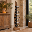 Teak Tree Root Wine Rack holding eight bottles of wine, standing beside a rustic wooden sideboard in a warm neutral room with a stone wall and a potted olive tree