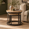 A round nesting side table with a light oak wood top and black metal frame, styled in a cosy living room next to a beige sofa with a vase and candle on top.